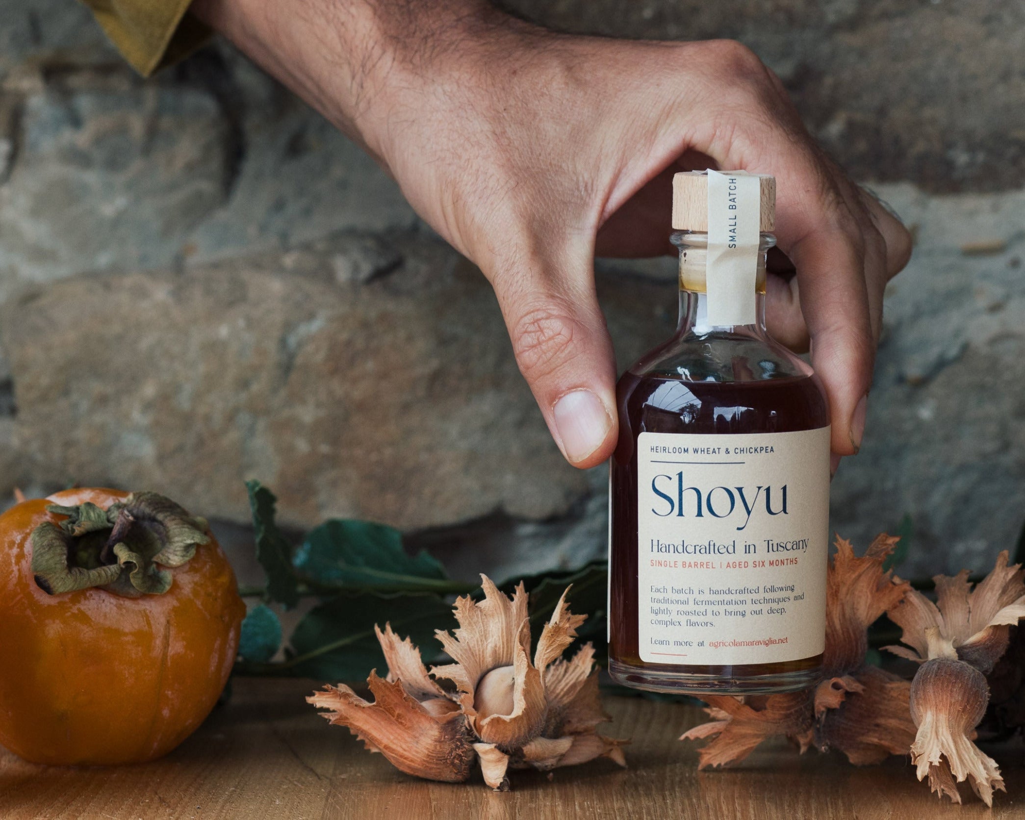 Hand holding a bottle labeled 'Shoyu' with a stone wall and persimmon fruit in the background