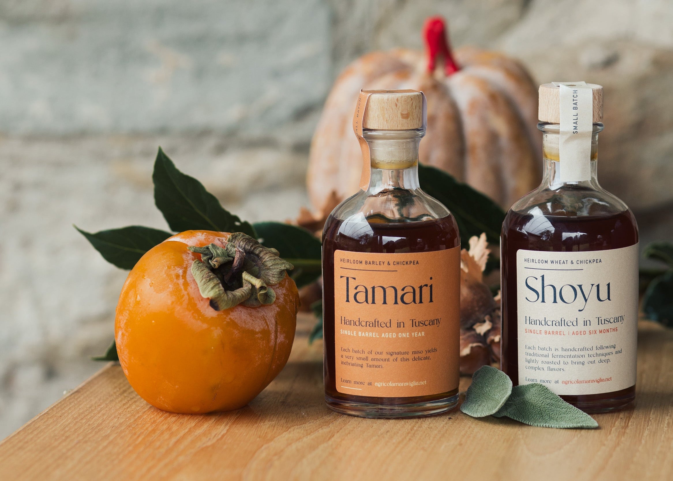 Two bottles of agricola maraviglia Shoyu and Tamari on a wooden surface with persimmons and leaves.