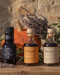 Three bottles of Maraviglia olive oil products—Olio, Tamari, and Shoyu—arranged on a rustic wooden table with a stone wall background, surrounded by lavender, olive branches, and a small pumpkin.